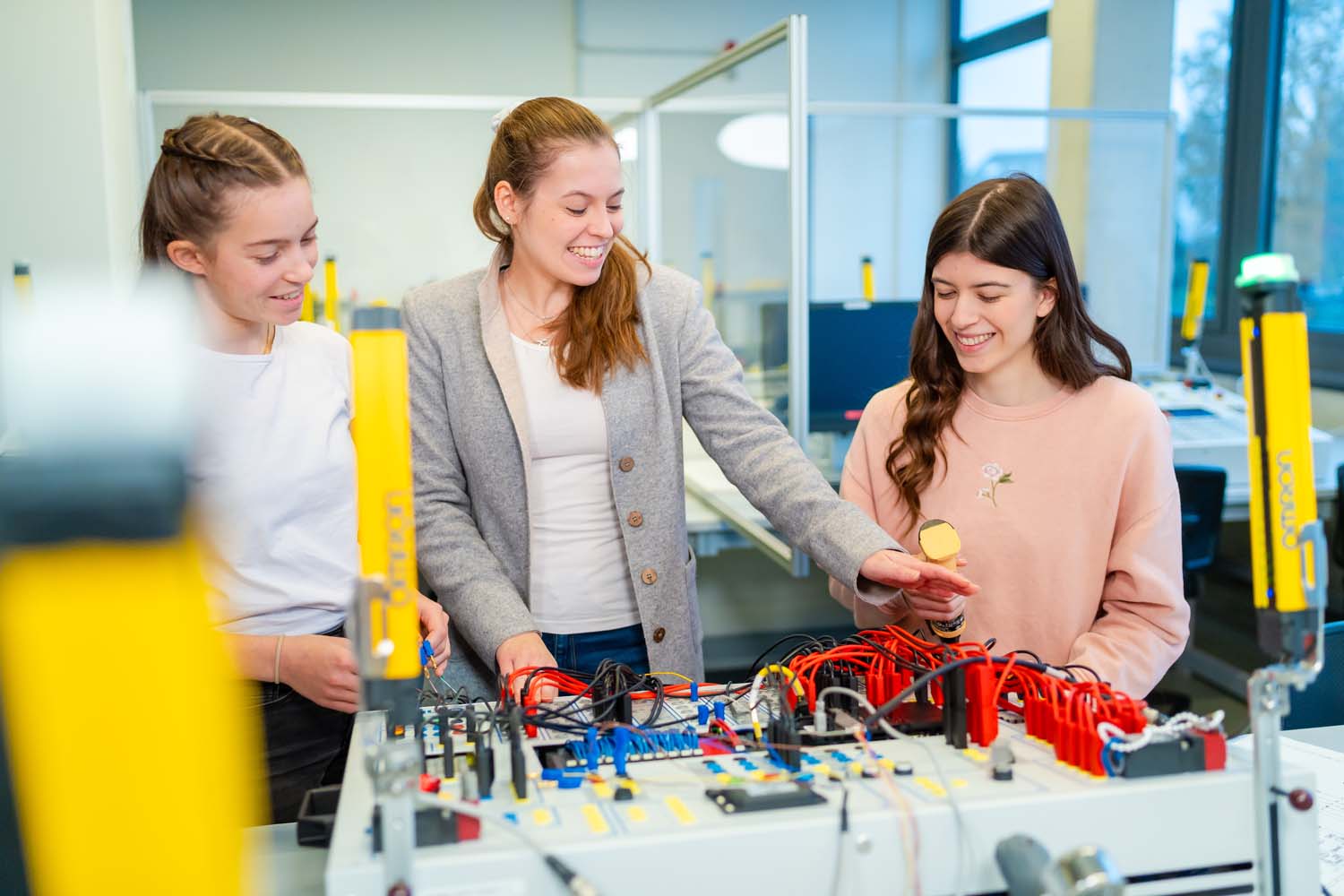 2 Studierende stehen an einem Schaltboard. 1 Lehrende erklärt die Aufgabe. Im Vordergund sieht man eine Lichtschranke