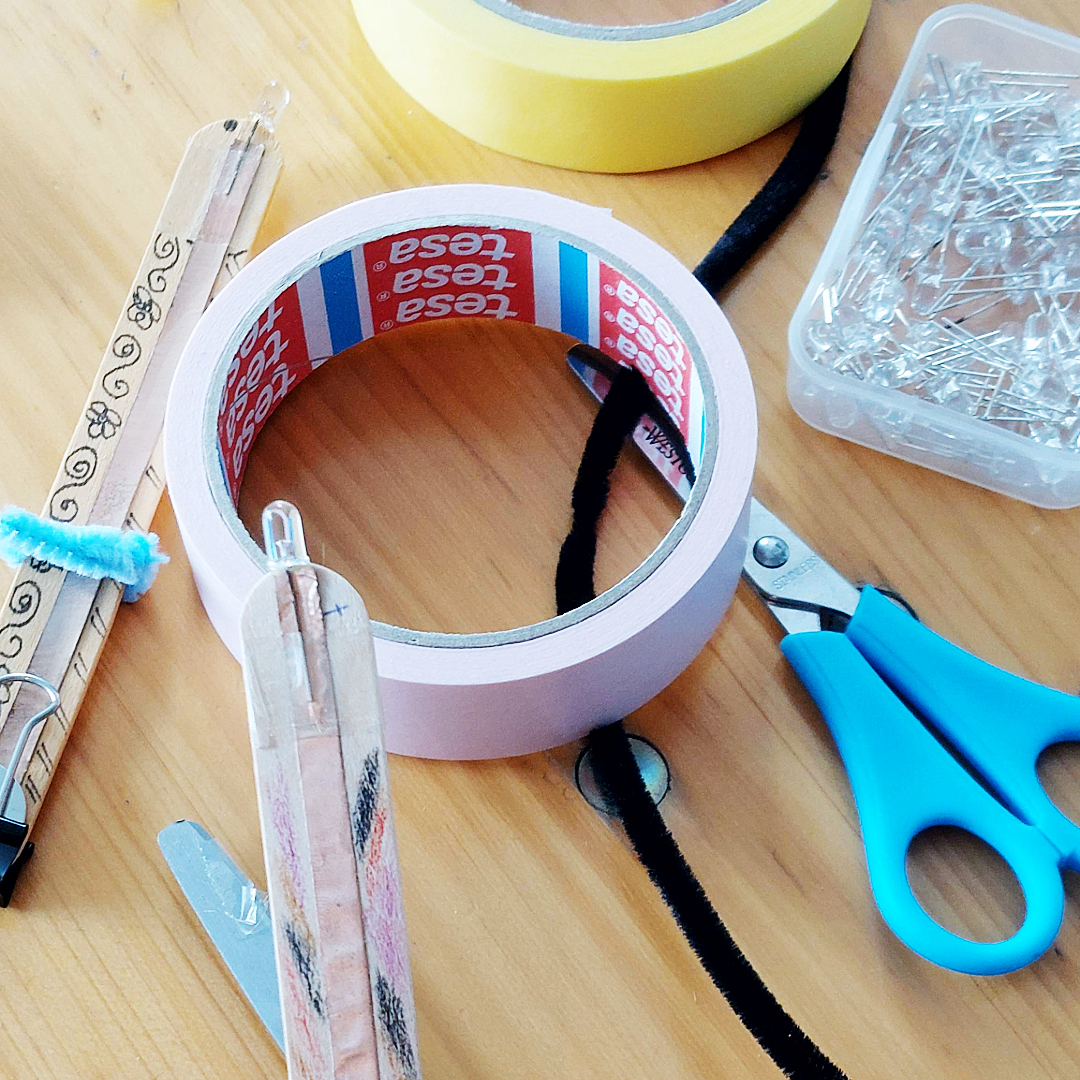Bastelmaterialien auf einem Holztisch: Sichtbar sind Klebeband, Schere, LED-Leuchten, Kupferband, einfache Büroklemmen und Holzstäbchen. Die Materialien werden für den Bau einfacher Taschenlampen verwendet.